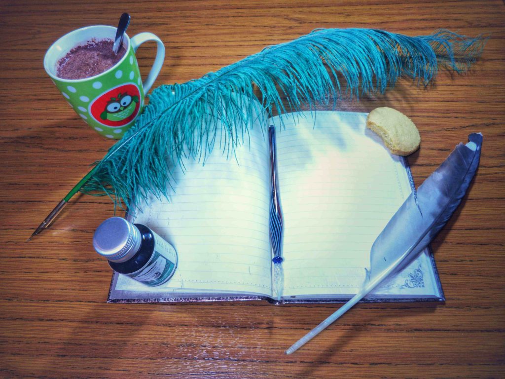 ästhetisiertes Foto des Schreibtischs der Autorin Alicia Hartung mit aufgeschlagenem Buch und altmodischen Schreibfedern und Tinte